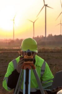 Surveyor in high-visibility gear and hard hat uses a theodolite on a tripod, with wind turbines at sunset in the background.